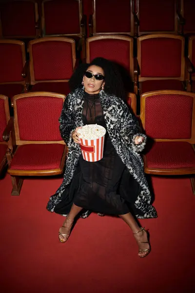 A stylish woman in a leopard print coat watches a movie, holding popcorn and reacting to the film. — Stock Photo