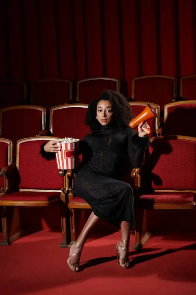 Una mujer con un vestido negro se sienta en un asiento de cine, sosteniendo palomitas de maíz y un megáfono. - foto de stock