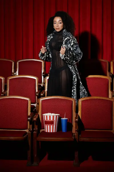 Uma mulher afro-americana elegante vestida com um casaco de pele elegante e vestido preto senta-se em um assento de cinema, assistindo a um filme. — Fotografia de Stock