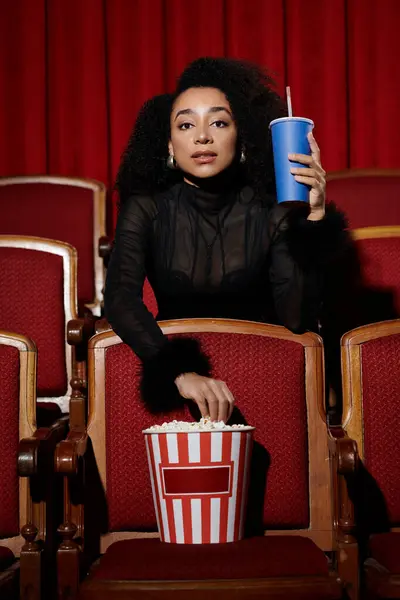 Una donna in maglia nera e acconciatura chic guarda un film in un teatro, immersa nel film con popcorn e un drink. — Foto stock