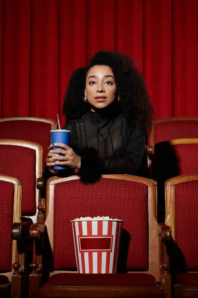 A stylish woman sits in a movie theater seat, holding a drink and watching the screen, her expression full of intrigue. — Stock Photo