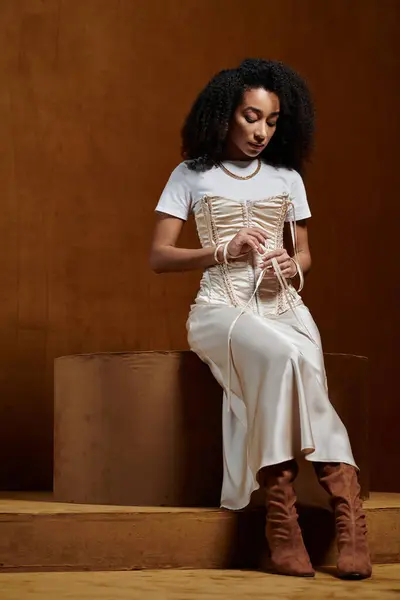 An African American woman in a white top and a cream satin skirt poses in a studio setting, adjusting the corset. — Stock Photo