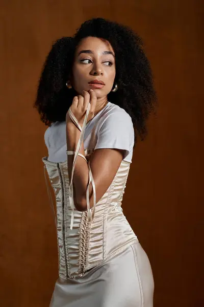 An elegant African American woman in a white top and cream corset poses confidently in a studio, dark curly hair framing her face. — Stock Photo
