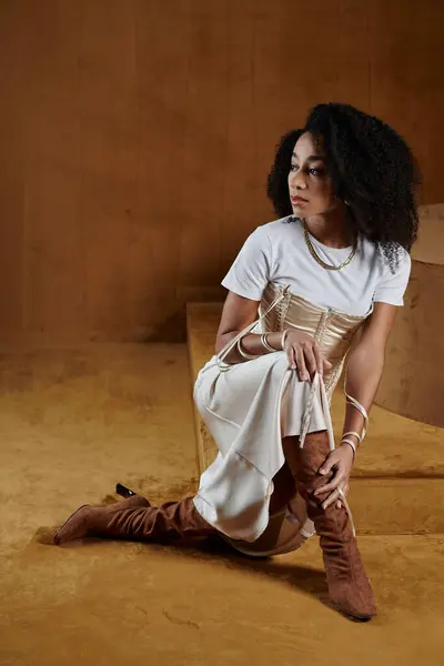 Une femme afro-américaine élégante dans une tenue tendance pose sur le sol d'un studio, son expression confiante et captivante. — Photo de stock