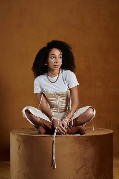 Une femme afro-américaine à la mode en t-shirt blanc et corset crème s'assoit sur une plate-forme marron, les jambes croisées. — Photo de stock