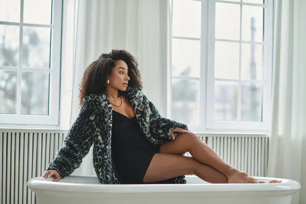 Una donna afroamericana alla moda in un vestito alla moda si siede in una vasca da bagno bianca in un ambiente da studio. — Foto stock