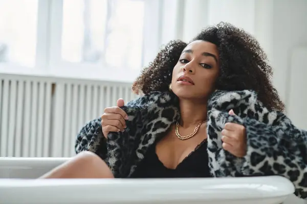 A woman with an afro in a fashionable leopard-print coat sits in a white bathtub and looks directly at the camera. — Stock Photo