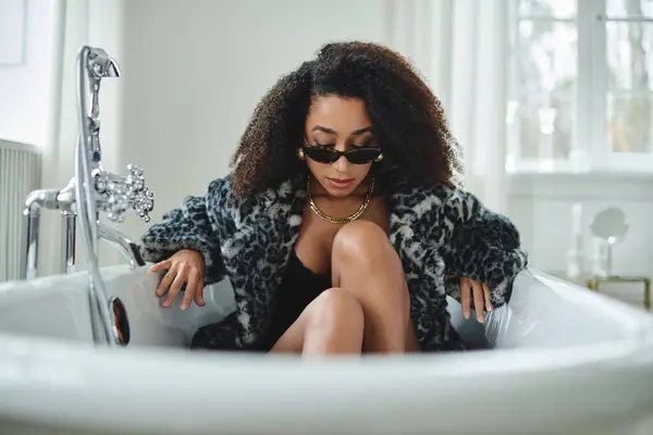 A stylish African American woman in a leopard print coat poses in a bathtub, with her legs crossed and her sunglasses resting on her nose. — Stock Photo