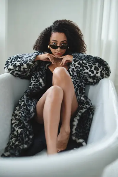 Una donna con un elegante cappotto leopardato si siede in una vasca da bagno bianca. Indossa occhiali da sole neri e un'espressione sicura. — Foto stock