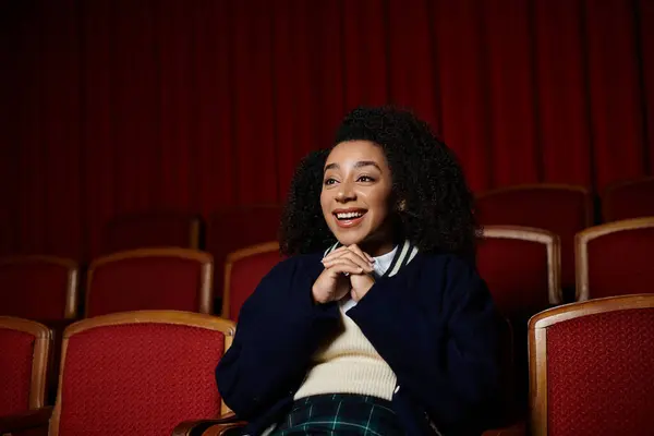 A young woman in casual clothes sits in a cinema seat, watching a movie with a surprised and delighted expression. — Stock Photo