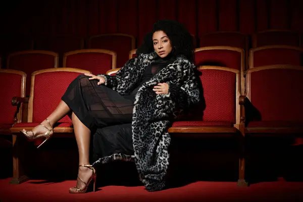 Stylish African American woman in leopard coat sits in cinema, reacting to movie on screen. — Stock Photo