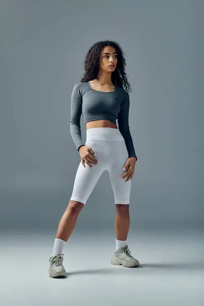 A young woman in a grey crop top and white shorts stands with one leg slightly bent, looking to the side. — Stock Photo