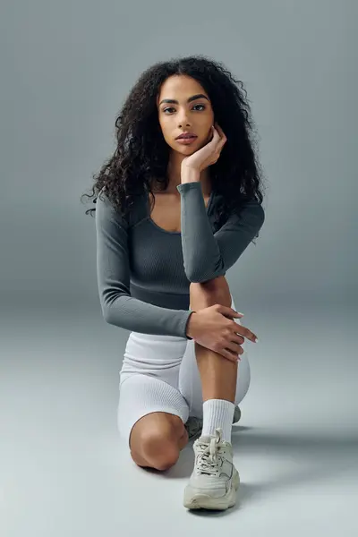 A young woman with curly hair sits on a grey studio floor, resting after a workout. — Stock Photo