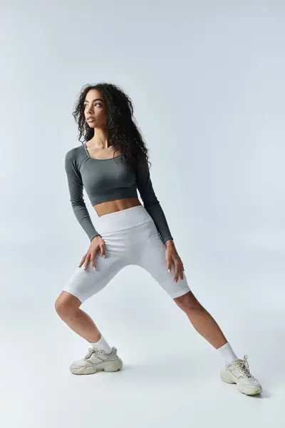 Una joven afroamericana con un top gris y pantalones cortos blancos se levanta con confianza, mostrando una poderosa postura de fitness. - foto de stock