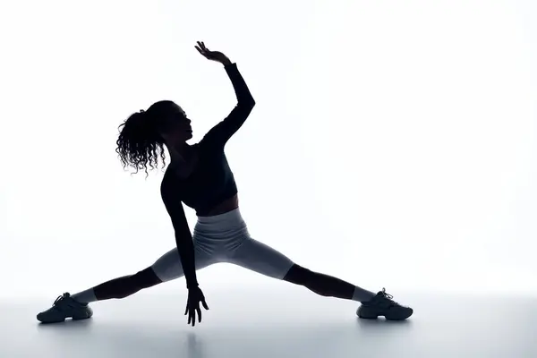 A young woman in athletic wear stretches gracefully, her silhouette standing out against a bright, white background. — Stock Photo