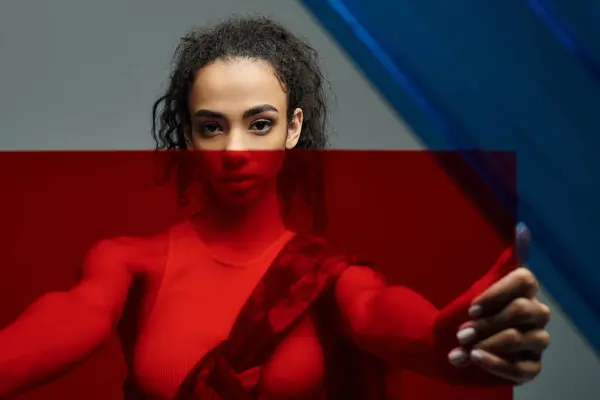 A black woman in red workout gear confidently holds a red glass panel against a gray backdrop with hints of blue, showing her strength. — Stock Photo