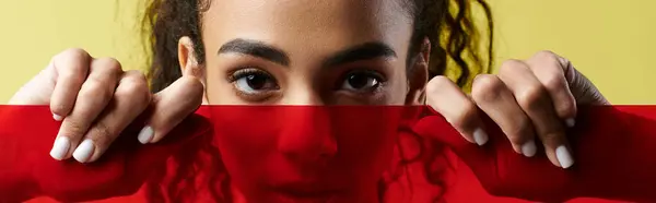 Молодая женщина с очаровательными глазами выглядывает из-за яркого красного стекла, излучая уверенность и стиль. — стоковое фото