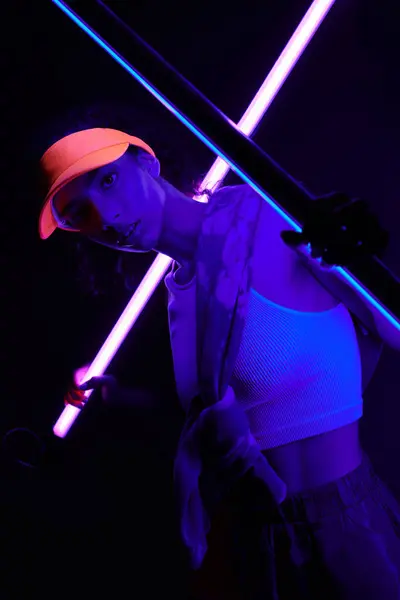 A young woman poses under vibrant neon lights, her orange cap casting a shadow across her face. — Stock Photo