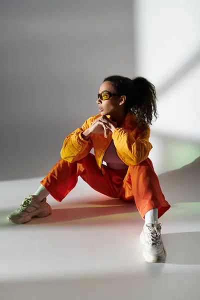 Un'elegante donna nera in giacca gialla, pantaloni arancioni, occhiali da sole e coda di cavallo guarda lateralmente. — Foto stock