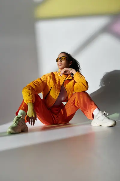 A young woman wearing a yellow jacket and orange pants sits on a white studio floor, posing with sunglasses. — Stock Photo