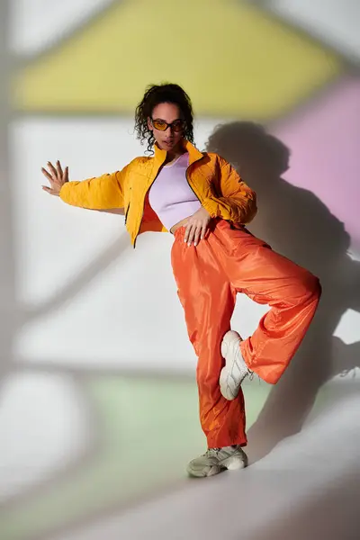 Una joven con chaqueta amarilla y pantalones naranjas posa con confianza sobre un fondo gris. — Stock Photo