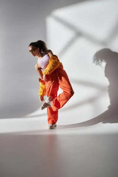 Una mujer afroamericana con gafas de sol, una chaqueta amarilla y pantalones naranjas posa sobre un fondo gris en un estudio. — Stock Photo