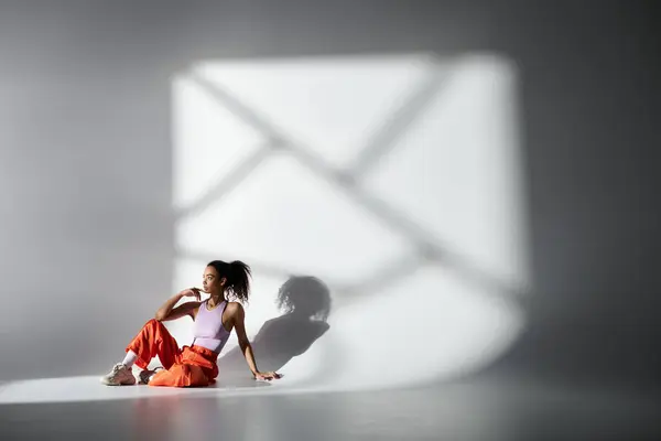 A young woman in orange pants and pink top sits in a studio, illuminated by a bright window casting a shadow on the grey wall. — Stock Photo