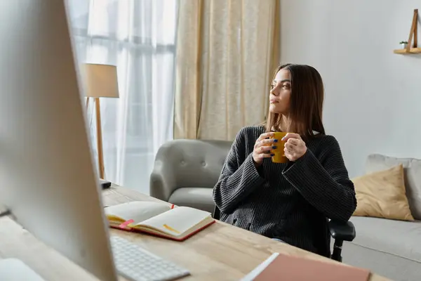 Una giovane donna, vestita con un maglione grigio, si siede a una scrivania nel suo ufficio, tenendo una tazza gialla, guardando attentamente fuori dalla finestra.. — Foto stock