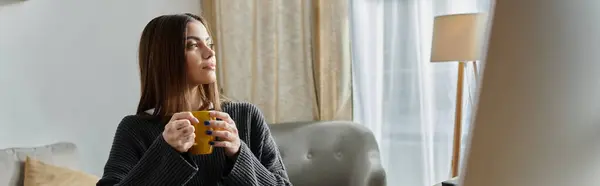 A young woman in a grey sweater sits thoughtfully in her home office, holding a warm drink. — Stock Photo