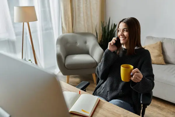 A young woman works on her computer while on a phone call, enjoying a cup of coffee. — Stock Photo