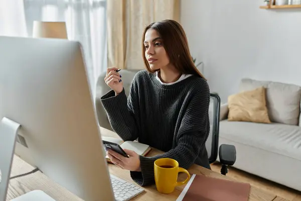 Woman in grey sweater, working at a desk in her home office on a computer with a coffee mug. — Stock Photo