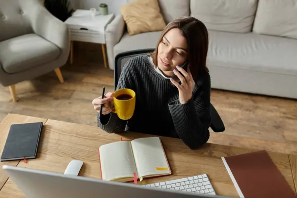 Una giovane donna si siede alla scrivania, lavora a distanza mentre tiene in mano una tazza di caffè e parla al telefono. — Stock Photo