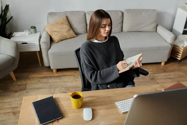 A young woman in a grey sweater sits at a desk in her home office, focused on writing in a notebook. — Stock Photo