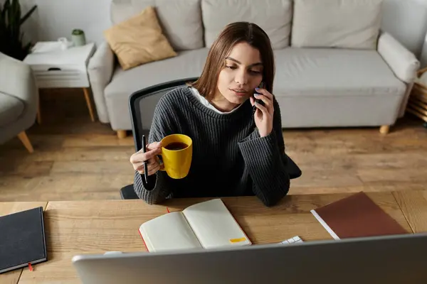 A young woman works from home, taking a break with a cup of tea while on a phone call. — Stock Photo