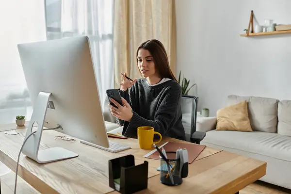 A young woman works at a desk in a home office, using her phone and a large monitor. — Stock Photo
