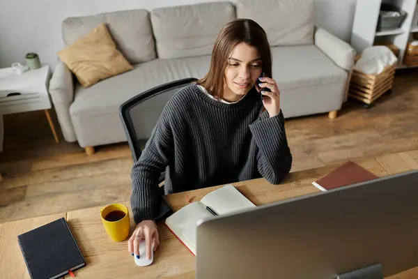 A young woman in a gray sweater sits at her desk, working on her computer while talking on the phone. — Stock Photo
