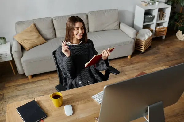A young woman in a grey sweater sits at her desk, working on her laptop and taking notes. — Stock Photo