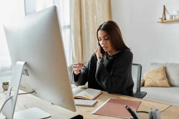 A young woman in a gray sweater sits at her desk, talking on the phone while working on her computer. — Stock Photo