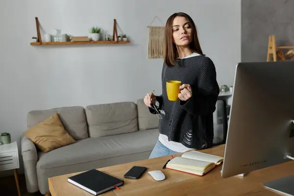 Una giovane donna con un maglione grigio lavora nel suo ufficio, tenendo in mano una tazza gialla di caffè. — Foto stock