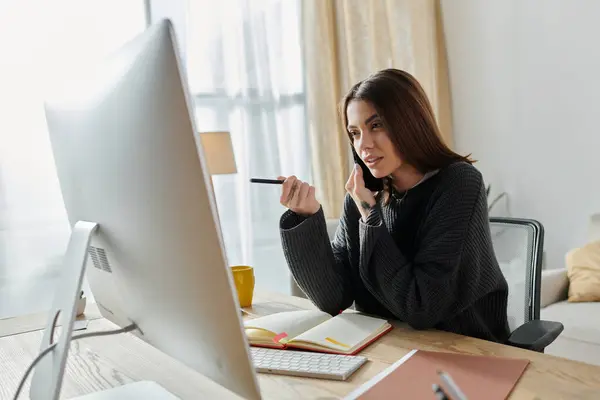 A young woman in a grey sweater sits at a desk in her home office, working on a computer while talking on the phone. — Stock Photo