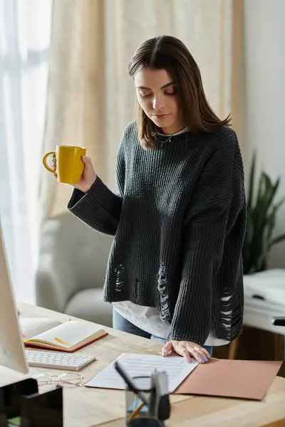 Young woman in grey sweater at home office, holding yellow mug, reviewing documents for social media content. — Stock Photo