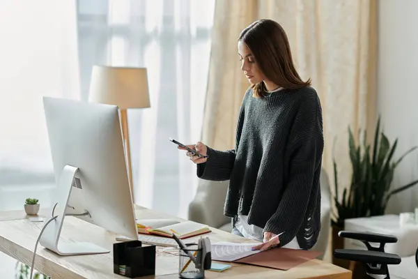 A young woman in a grey sweater works at her home office desk, looking at her phone and holding paperwork. — Stock Photo
