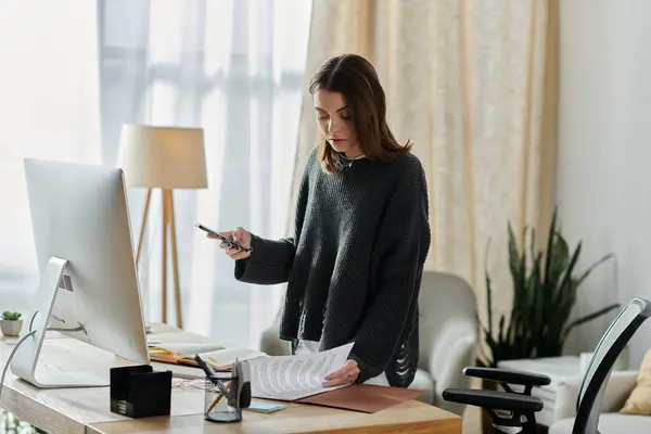 Une jeune femme dans un pull gris se tient à son bureau, travaillant sur son ordinateur et son téléphone. — Photo de stock