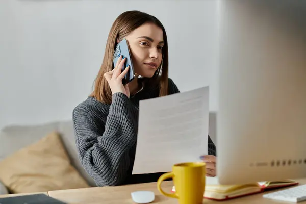 A young woman in a grey sweater works on a project from home while taking a phone call. — Stock Photo