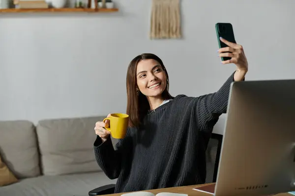 Una mujer joven se sienta en un escritorio en su oficina, tomando una selfie con su teléfono mientras sostiene una taza en su otra mano. - foto de stock