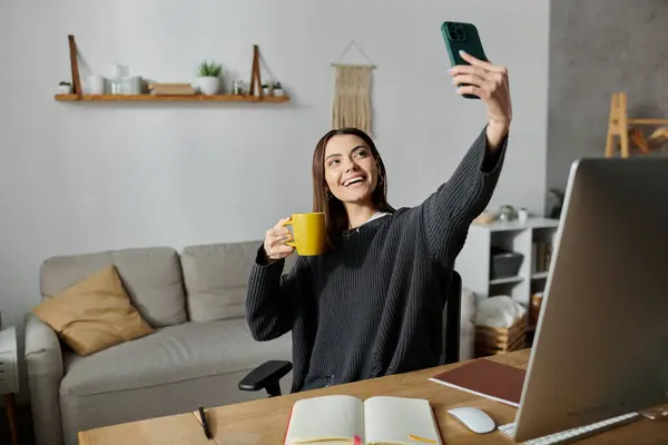 Uma mulher de camisola cinza tira uma selfie enquanto trabalha em casa. Ela está segurando uma caneca amarela e sorrindo. — Fotografia de Stock