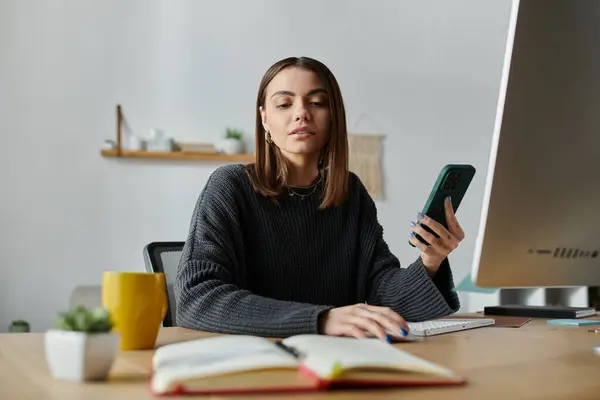 A young woman in a grey sweater sits at a desk in her home office, working on her laptop while holding a phone in her hand. — Stock Photo