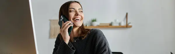 Uma jovem mulher em uma camisola cinza senta-se em seu escritório em casa, falando em seu telefone e sorrindo. — Fotografia de Stock