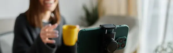A woman in grey records a video at home, smiling, gesturing, and holding coffee for social media. — Stock Photo