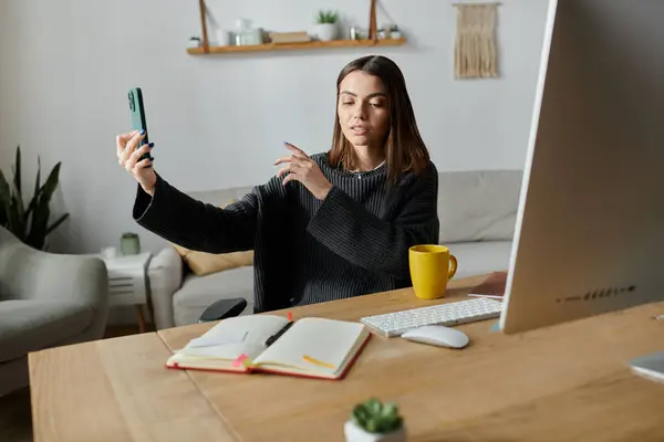 Una mujer joven con un suéter gris filma contenido en su teléfono mientras está sentada en un escritorio en su oficina en casa. - foto de stock